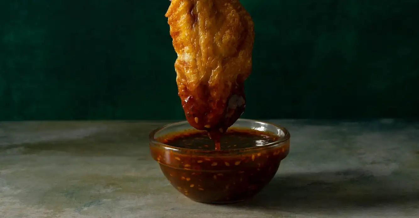 Golden-brown fried chicken wing being dipped into a glass bowl of glossy dark red spicy gochu sauce