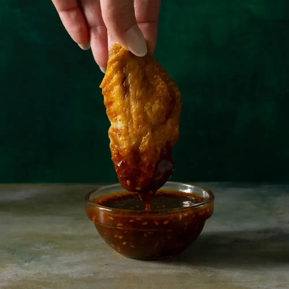 A golden-brown, crispy fried chicken wing being dipped into a bowl of spicy red sauce.