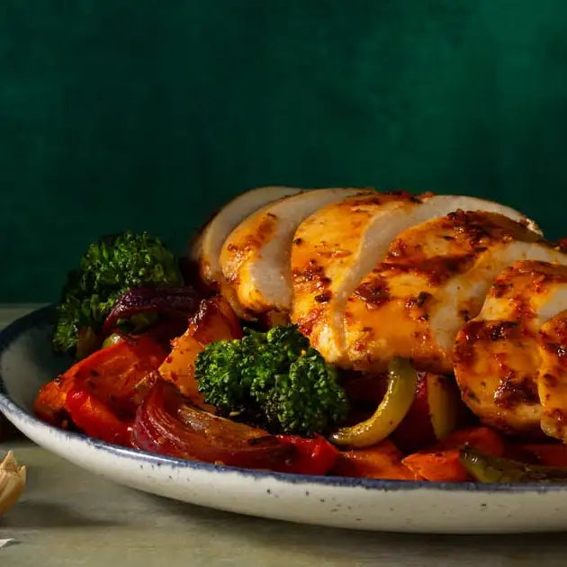 A plate of grilled chicken breast slices with a golden-brown, slightly charred surface, served alongside vibrant roasted vegetables including broccoli, red peppers, and onions on a speckled white ceramic dish.