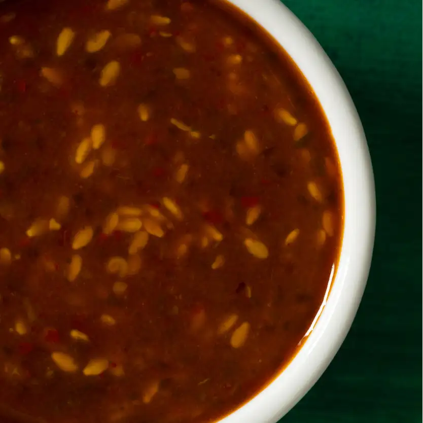 A rich, dark brown sauce filled with golden sesame seeds and flecks of red chili in a white ceramic bowl.
