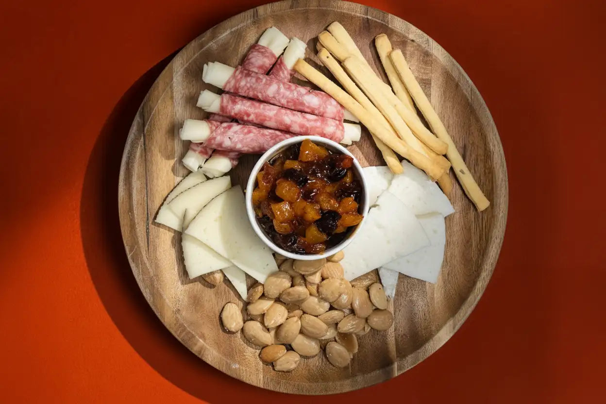 A round wooden platter holding an assortment of cheeses, cured meats, breadsticks, nuts, and a small bowl of fruit compote.