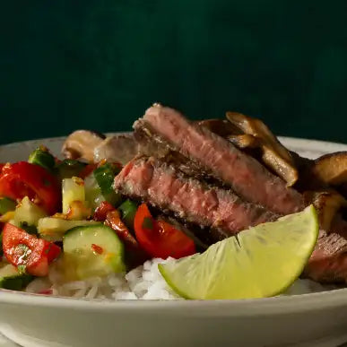 A savory beef bowl with sliced medium-rare steak, sautéed mushrooms, and a colorful medley of diced tomatoes, cucumbers, and green peppers atop white rice, garnished with a bright lime wedge.