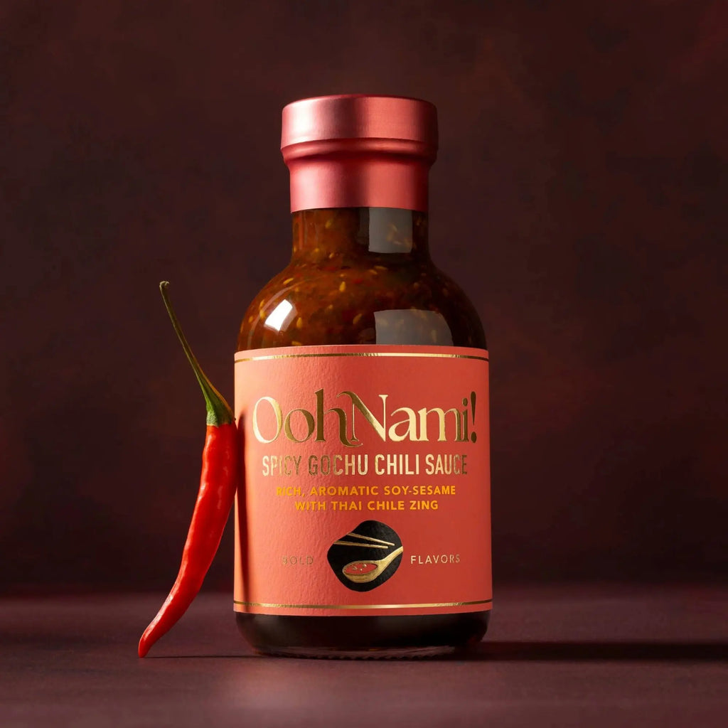 A bottle of ooh nami spicy gochu chili sauce with a reddish-pink label and metallic pink cap featuring soy-sesame flavor