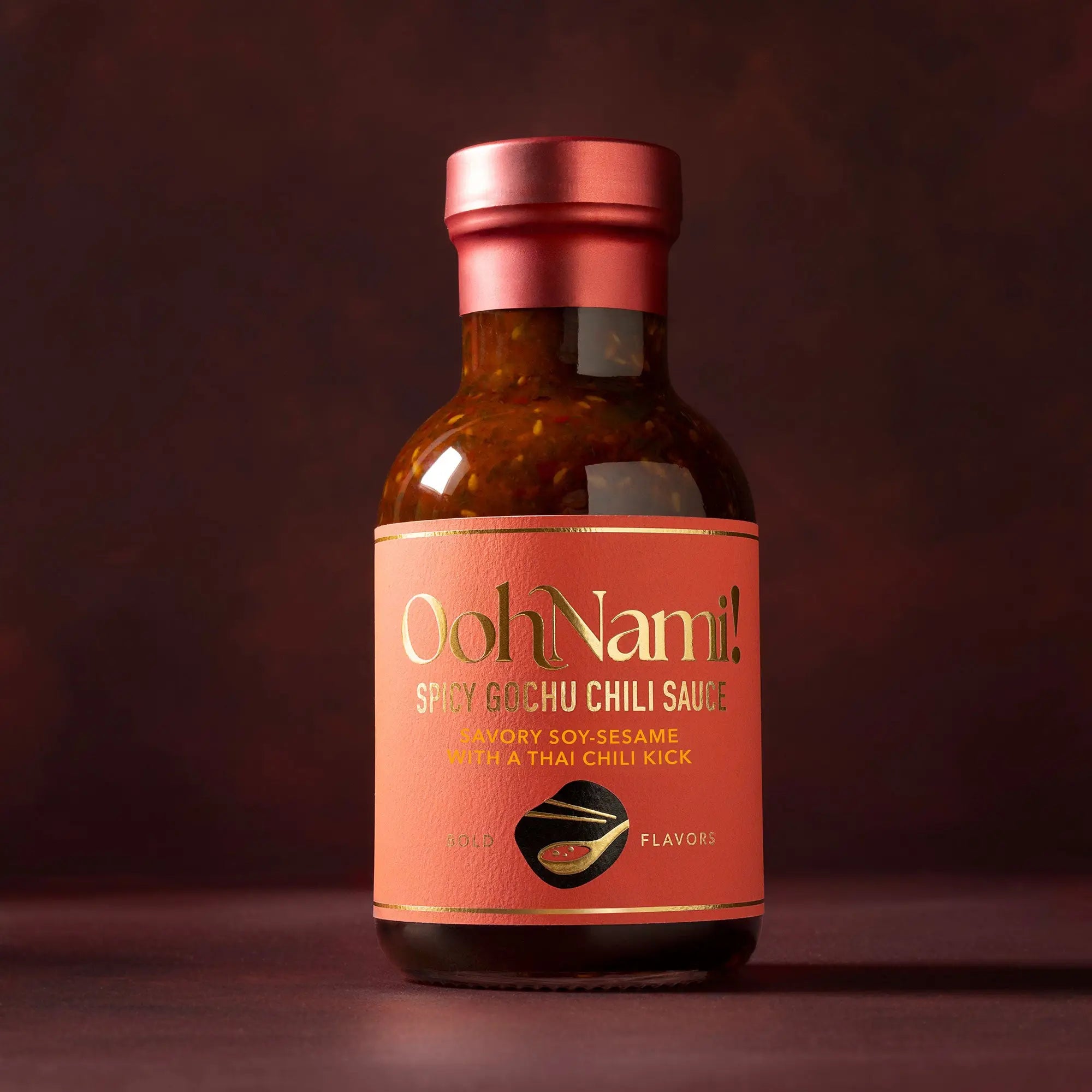 A sleek amber glass bottle with a metallic rose gold cap and label for ooh nami spicy gochu chili sauce