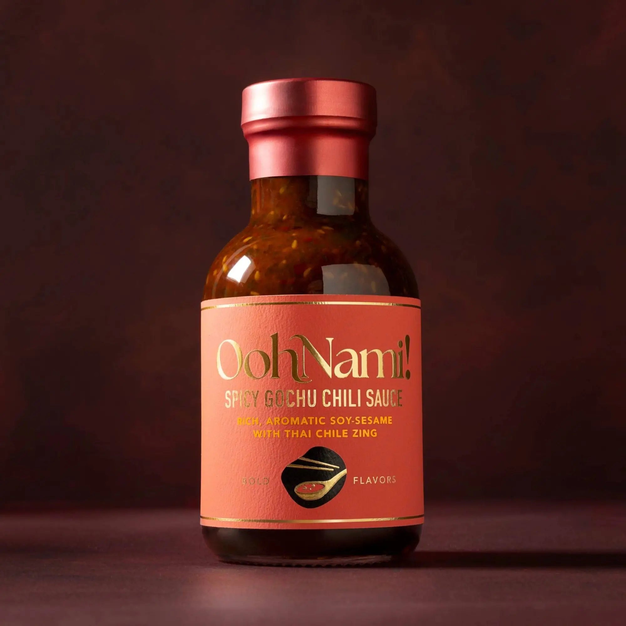 A sleek amber glass bottle with a metallic rose gold cap and label for ooh nami spicy gochu chili sauce