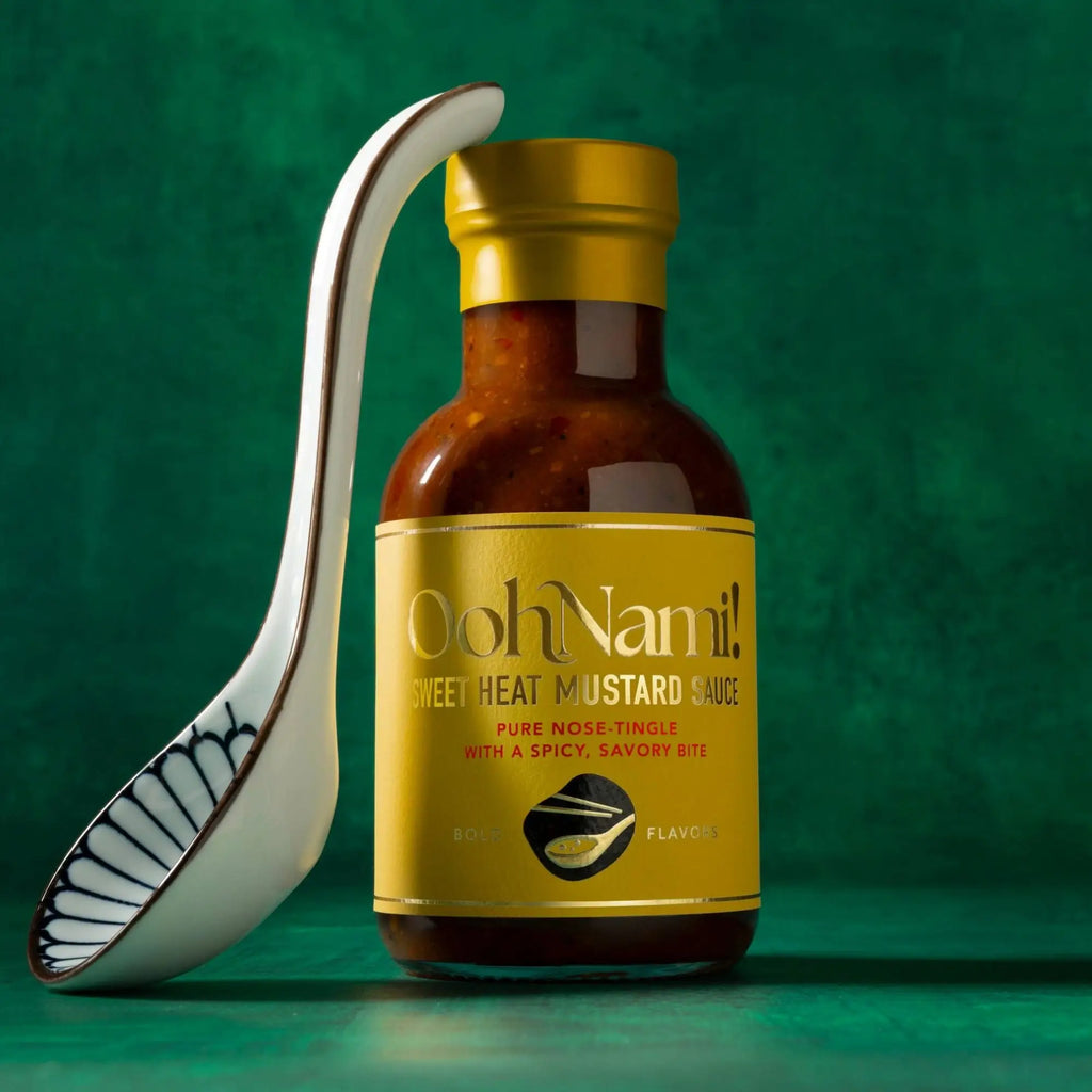 A sleek amber glass bottle of oohnami sweet heat mustard sauce with a gold cap and a white ceramic spoon with black patterns