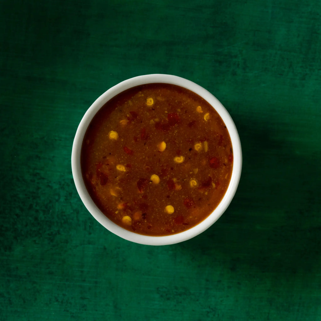 A white ceramic bowl filled with chunky reddish-brown sweet heat mustard sauce with corn and red pepper flakes