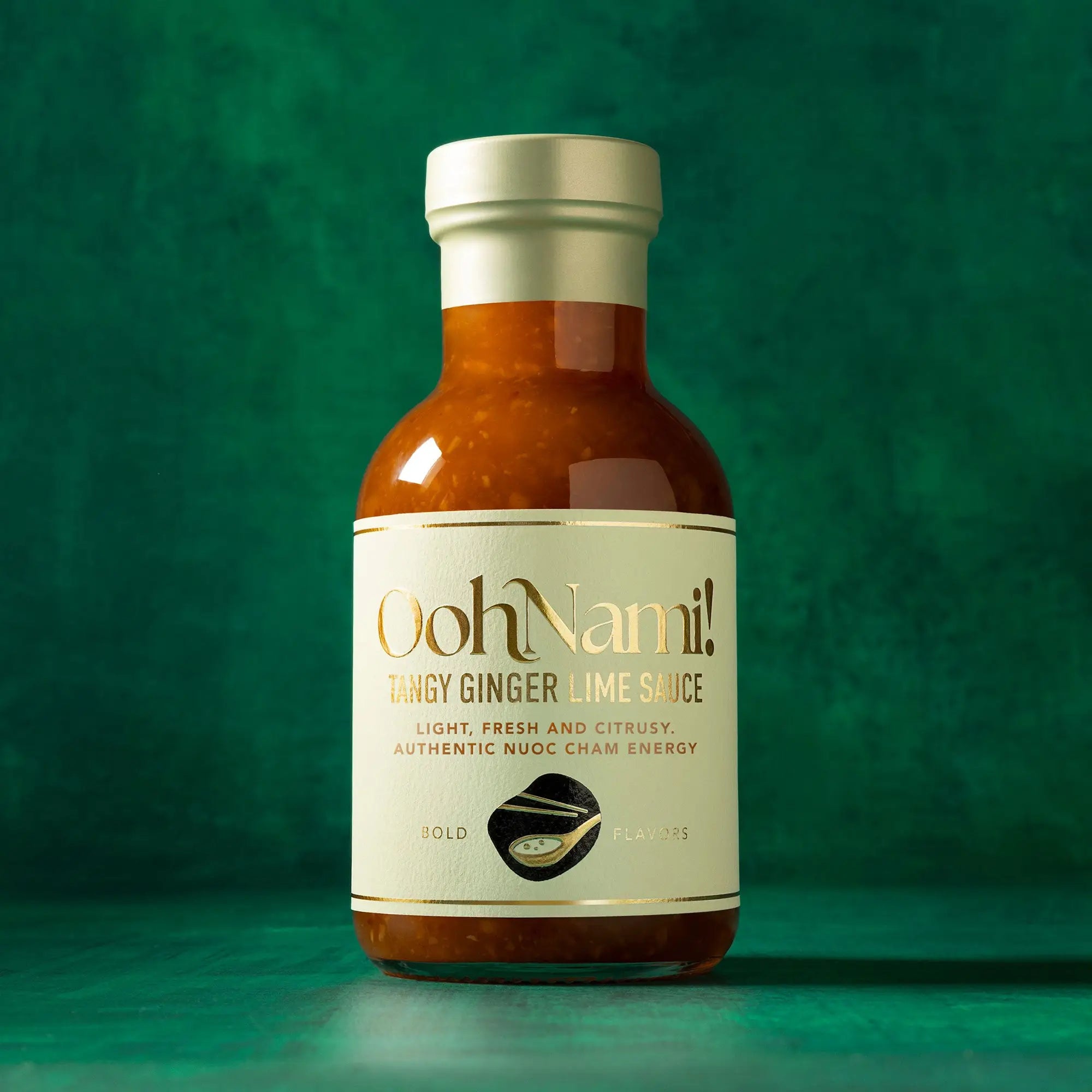 A glass bottle of Oohnami tangy ginger lime sauce with white label and gold lettering filled with orange-red sauce