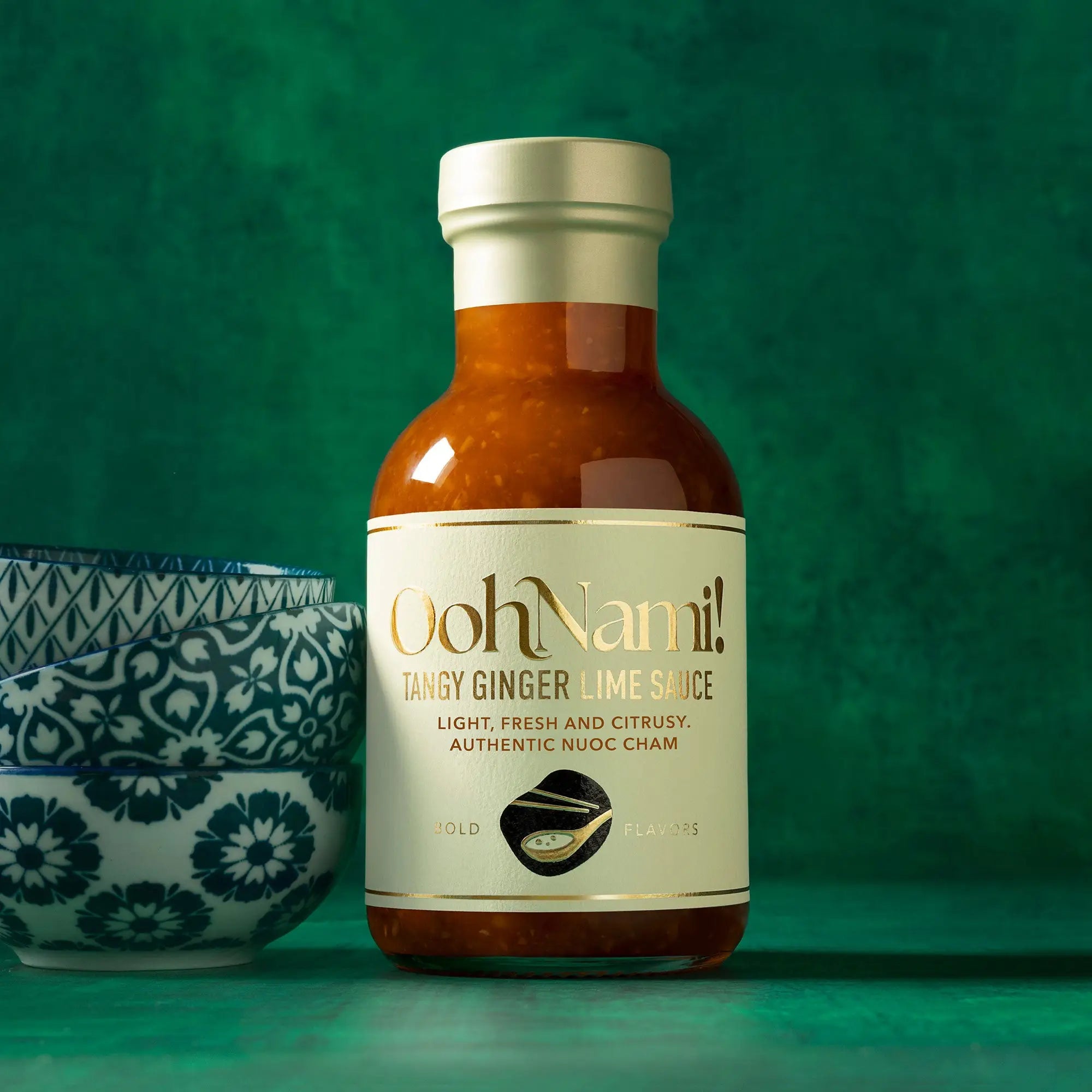 A glass bottle of Oohnami tangy ginger lime sauce with a metallic silver cap and cream label with gold lettering