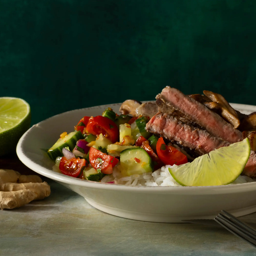 A white ceramic bowl with vibrant salad topped with sliced steak and lime wedges paired with tangy ginger lime sauce