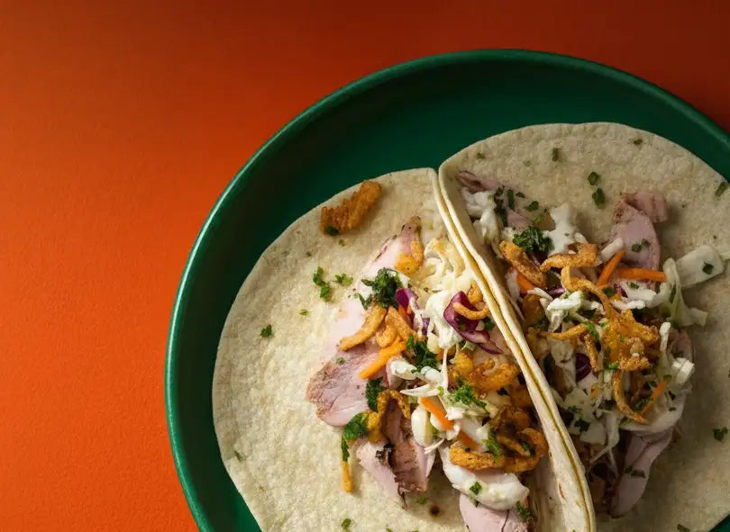 Two soft tortillas filled with sliced meat, shredded cabbage slaw, crispy fried onions, and fresh herbs are served on a green plate.