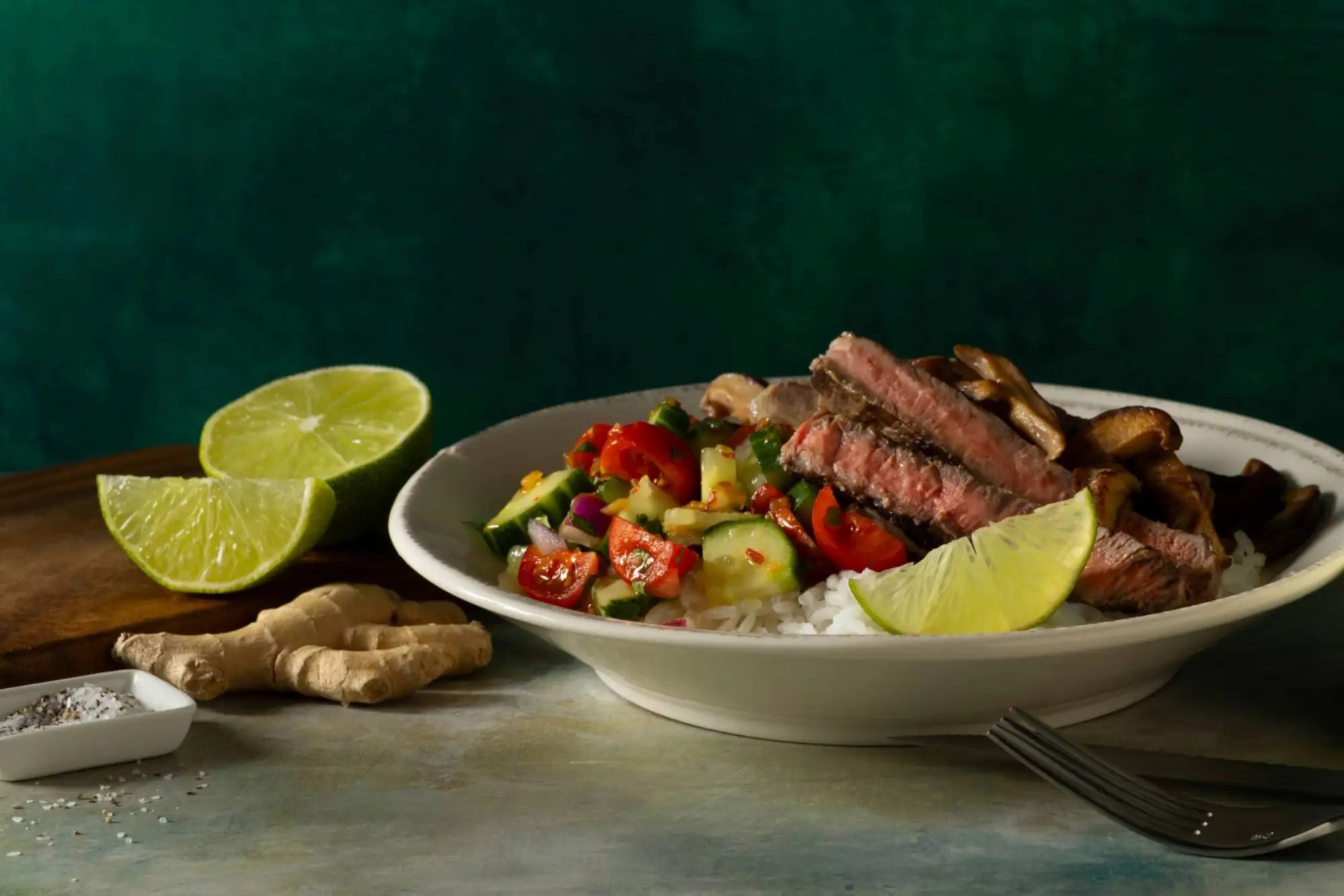 A white ceramic bowl filled with a colorful medley of sliced steak, rice, diced vegetables, and garnished with lime wedges.