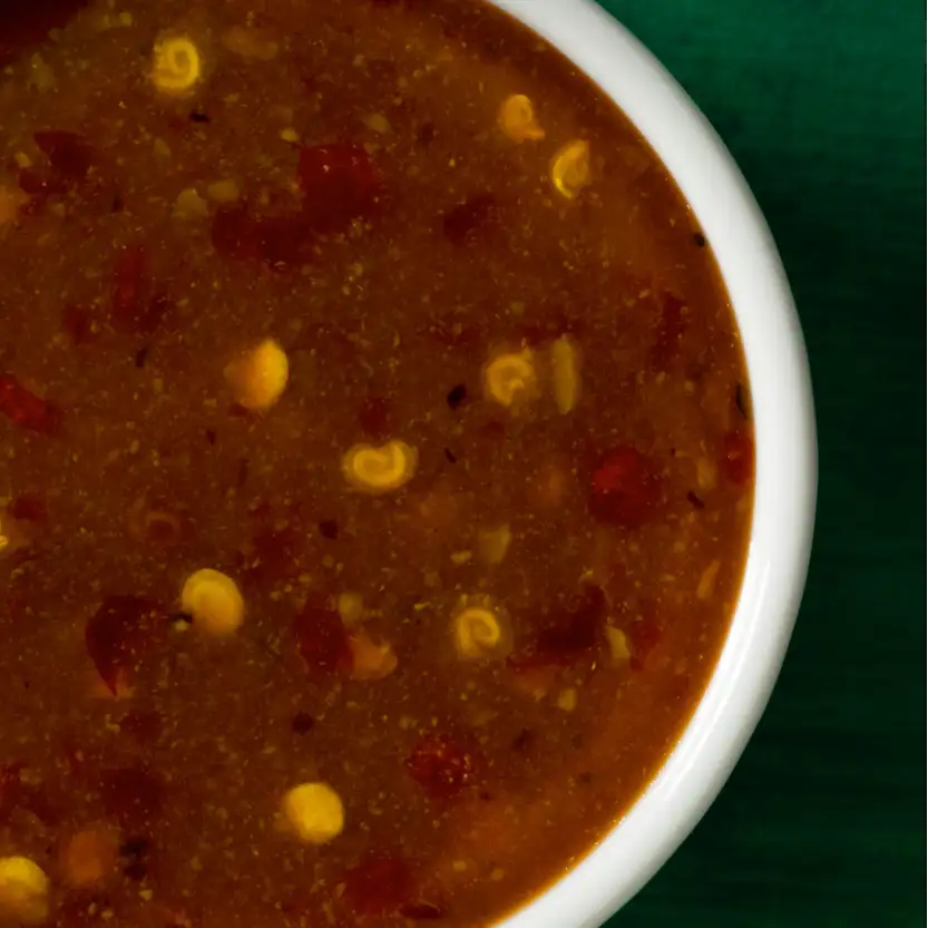 A white ceramic bowl filled with a rich, reddish-brown sauce dotted with yellow corn kernels and red chili pieces.