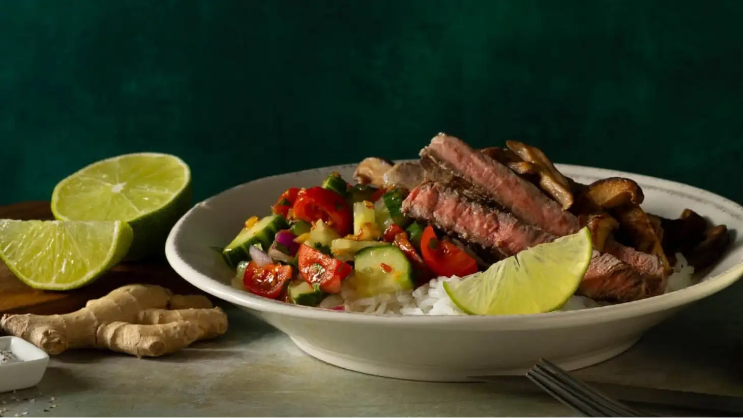 A white ceramic bowl filled with a vibrant salad of diced tomatoes, cucumbers, and herbs atop rice, garnished with sliced steak and lime wedges.