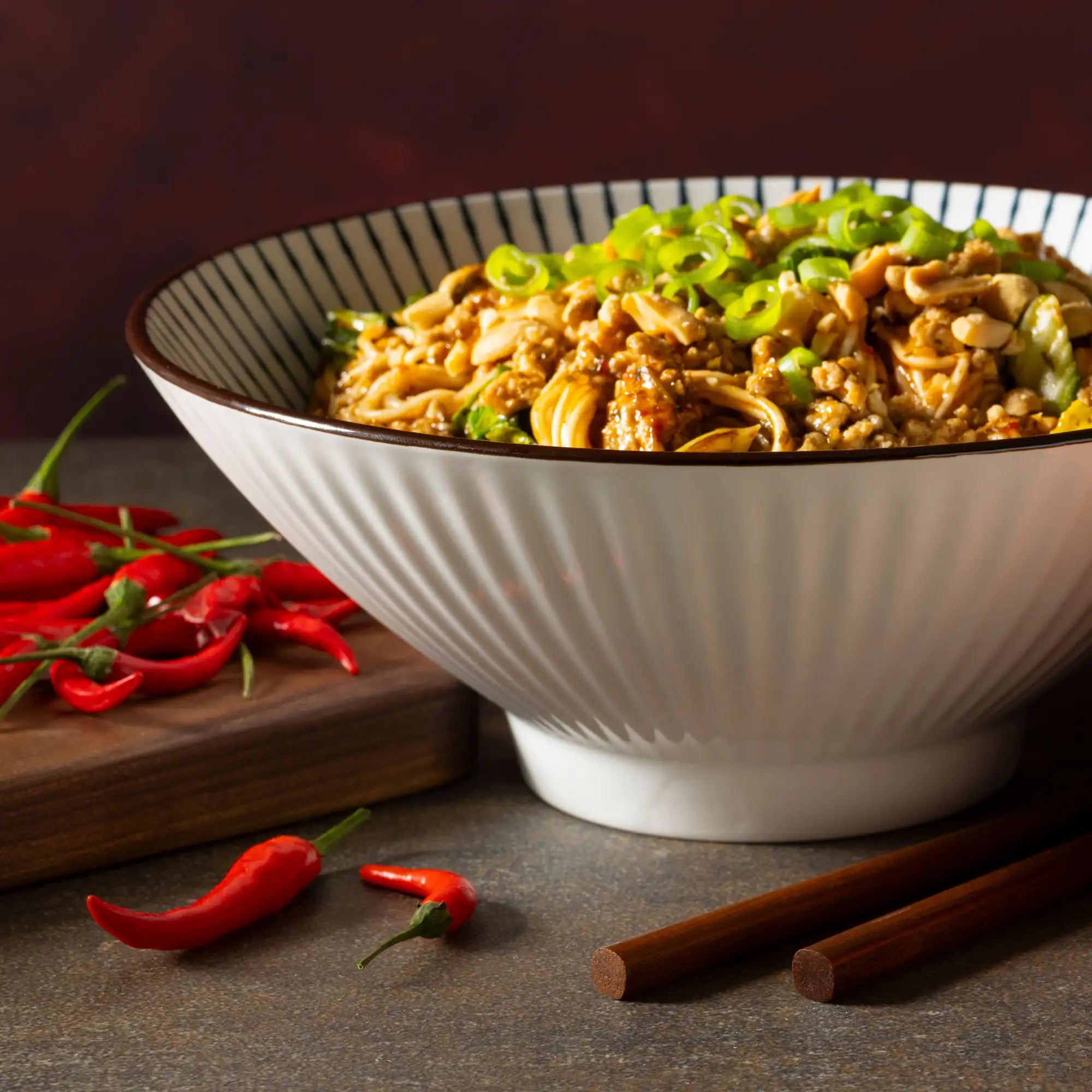 A white ceramic bowl with a ribbed exterior and black-striped rim, filled with stir-fried noodles topped with green onions.