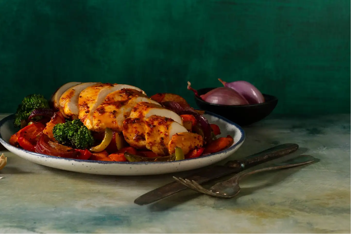 A white ceramic plate with a blue rim, holding sliced grilled chicken breast over a bed of roasted vegetables.